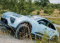 A light blue Porsche Macan is driving off-road on an uneven, grassy terrain with trees and shrubs in the background.