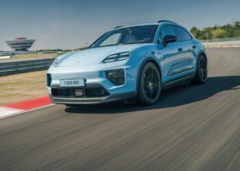A blue Porsche Macan SUV with “1,000,000” on the license plate speeds along a racetrack, celebrating the millionth Macan, with blurred motion and a building visible in the background.