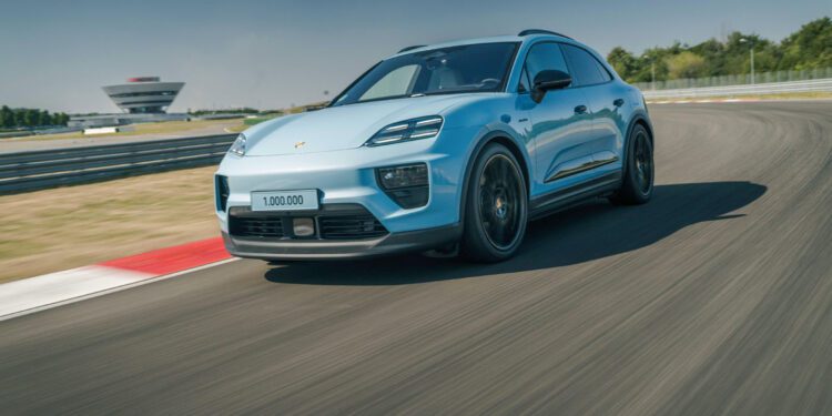 A blue Porsche Macan SUV with “1,000,000” on the license plate speeds along a racetrack, celebrating the millionth Macan, with blurred motion and a building visible in the background.