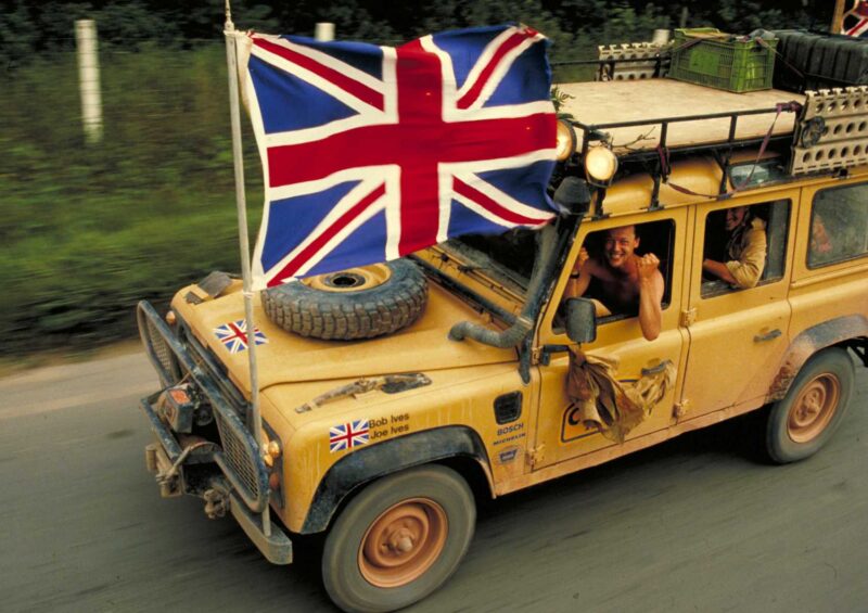 A yellow Land Rover Classic with a large Union Jack flag, roof cargo, and stickers drives on the road as smiling passengers wave—evoking the spirit of 10 historic expedition icons set to showcase at the 2025 Pebble Beach Concours d'Elegance.