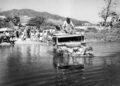 A group of people watch as a Land Rover Classic with passengers on top crosses a shallow river, hills and trees in the background—evoking the spirit of expedition icons set to be showcased at the 2025 Pebble Beach Concours d'Elegance.