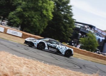 A Zenvo Aurora sports car with geometric camouflage and "Aurora" branding makes its global debut on a racetrack bordered by hay bales, with trees and a multi-level building in the background.