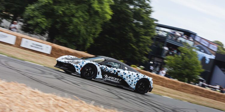 A Zenvo Aurora sports car with geometric camouflage and "Aurora" branding makes its global debut on a racetrack bordered by hay bales, with trees and a multi-level building in the background.