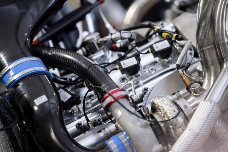 Close-up view of a modern V8 Twin-Turbo engine with visible hoses, wires, and metal components, including a black air intake pipe with blue bands and a silver turbocharger—engineered for Genesis Magma Racing hypercar performance.