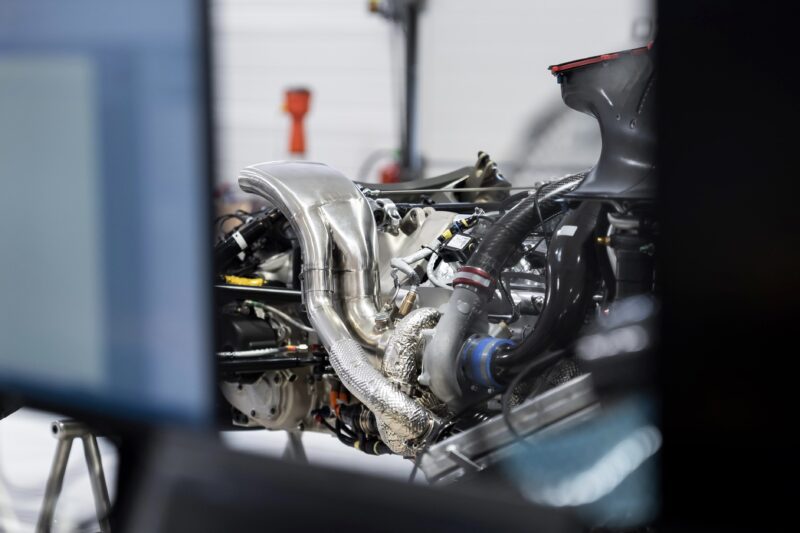 A close-up view of a complex GMR‑001 Hypercar engine with visible metal pipes, wiring, and V8 Twin-Turbo components in a workshop setting.