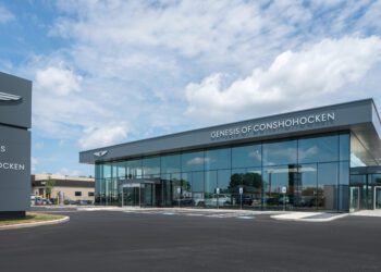 Modern car dealership building with large windows and a sign reading "Genesis of Conshohocken" under a partly cloudy sky.
