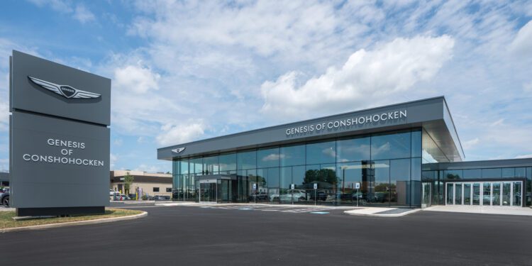Modern car dealership building with large windows and a sign reading "Genesis of Conshohocken" under a partly cloudy sky.