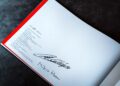 Open book displaying printed text in multiple languages and two large handwritten signatures on a blank white page, evoking memories of Lanzante’s iconic McLaren F1 GTR at Goodwood.