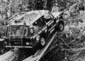 A Land Rover Classic with expedition markings crosses a narrow metal plank bridge in a dense jungle; a shirtless person stands nearby on the roof, echoing the spirit of the 10 Historic Expedition Icons set for the 2025 Pebble Beach Concours d'Elegance.