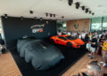 A crowd gathers around a covered car and an orange Lamborghini Temerario on display at a Lamborghini event indoors, reminiscent of the excitement found at Goodwood.