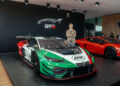 A man in a suit stands with arms crossed beside a Lamborghini Huracán GT3 EVO2 Temerario race car, displayed on a platform at Goodwood's prestigious auto show.