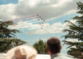 Seven planes fly in formation, leaving red, white, and blue smoke trails above Goodwood, as two people watch from below on a partly cloudy day.
