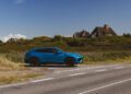 A bright blue SUV is parked on the side of a rural road with grass, shrubs, and thatched-roof houses in the background under a clear sky.