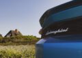 Close-up of the rear end of a blue Lamborghini car with a house and greenery blurred in the background under a clear sky.