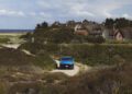 A blue car drives on a narrow path through grass and shrubs, with thatched-roof houses and the sea visible in the background under a partly cloudy sky.