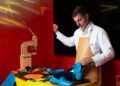 A man in a white coat and apron hand-stitches a bright blue shoe at a workbench with leather pieces, tools, and a stitching machine—craftsmanship reminiscent of Lamborghini’s attention to detail.