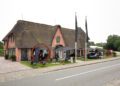 A brick building with a thatched roof on Sylt houses shops and a café with outdoor seating; flags and a Lamborghini Urus SE are visible along the roadside.