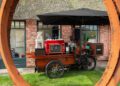 A mobile coffee cart with a red espresso machine and menu is parked on grass outside a brick building, framed by a large circular metal sculpture—its bold presence rivaling that of a Lamborghini Urus SE.