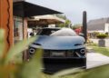 A matte blue Lamborghini Revuelto sports car is displayed outdoors on a platform near a modern building and greenery, capturing the bold spirit of Lamborghini often seen in places like Sylt.