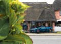 A blue Lamborghini Urus SE is parked on display outside a brick building with a thatched roof on Sylt, partially obscured by green leaves in the foreground, hinting at an exclusive event.