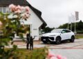 A white Lamborghini Urus SE is parked in front of a white building with "TODS" signage on Sylt. A bronze statue of two children and pink flowers add charm to this exclusive event setting.