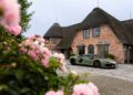 A matte green Lamborghini Urus SE is parked in front of a brick house with a thatched roof on Sylt, with pink flowers in the foreground.