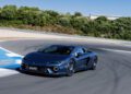 A blue Lamborghini Temerario makes a dynamic debut as it speeds around a curve on the track, with trees and a building in the background under a clear sky.