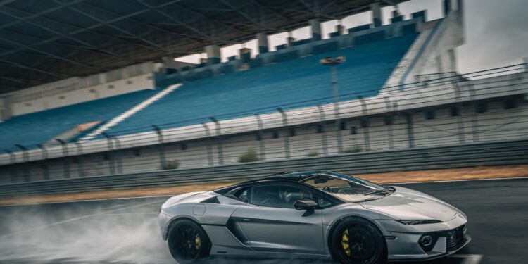 A silver Lamborghini Temerario makes a dynamic debut as it speeds on track, sending water spray flying near empty blue stadium seats under a large canopy.