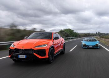Two cars, an orange SUV and a blue Lamborghini sports car, drive side-by-side on a multi-lane highway under a cloudy sky.