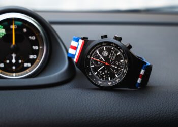 A black Chronograph 1 wristwatch with a red, white, and blue striped fabric strap, designed by Porsche Design, displayed on a car dashboard next to a round speedometer—perfect for PCA enthusiasts.