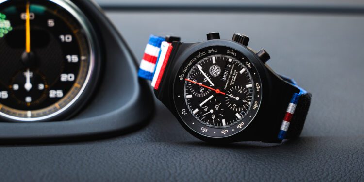 A black Chronograph 1 wristwatch with a red, white, and blue striped fabric strap, designed by Porsche Design, displayed on a car dashboard next to a round speedometer—perfect for PCA enthusiasts.