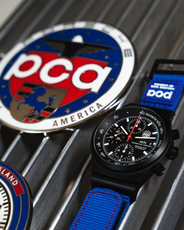 A black Porsche Design Chronograph 1 wristwatch with a blue strap sits on a metal surface next to a round badge with "PCA" and "America" text.