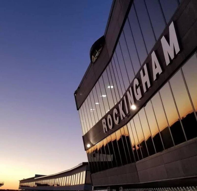A modern building with large windows reflects the sunset sky. The word "ROCKINGHAM" is prominently displayed on the side, highlighting its close proximity to Rockingham Speedway—an ultimate playground for racing enthusiasts.