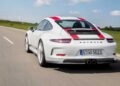 A 2016 Porsche 911 R, white with red racing stripes, drives on a rural road surrounded by green fields and a partly cloudy sky.