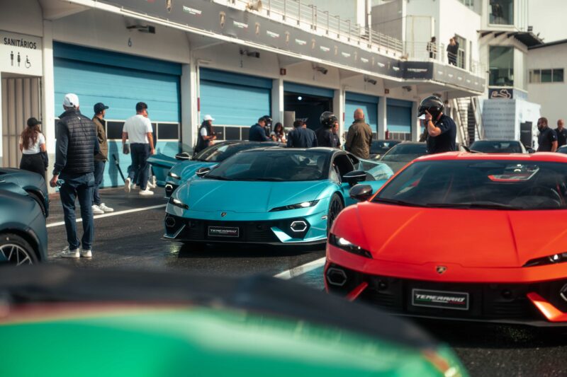 A group of people stand near several Lamborghini sports cars, including the Lamborghini Temerario and V12 Revuelto, parked outside garages at a racetrack event for a thrilling track review.