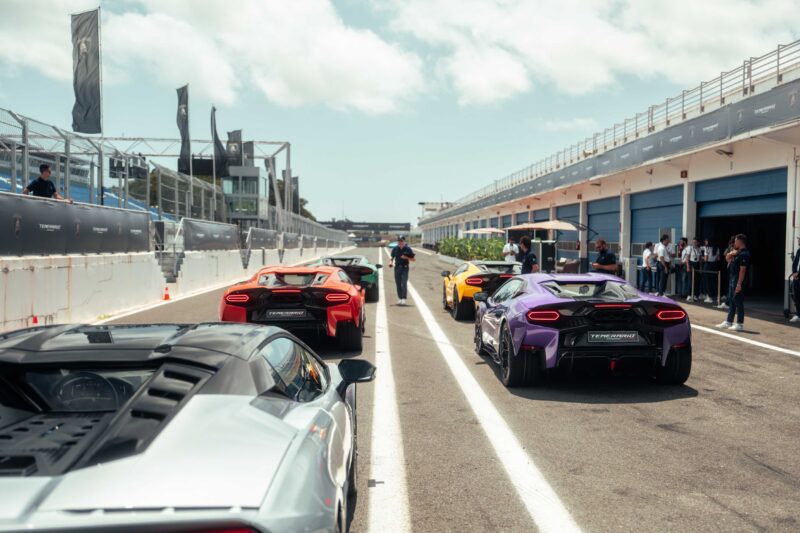 Several luxury sports cars, including the V12 Revuelto and Lamborghini Temerario, are parked on a racetrack pit lane for a track review, with people standing nearby and garages in the background under a partly cloudy sky.