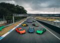 Seven brightly colored Lamborghini sports cars, including the Lamborghini Temerario and V12 Revuelto, are parked on a racetrack under cloudy skies, viewed from above—a perfect scene for a thrilling track review.