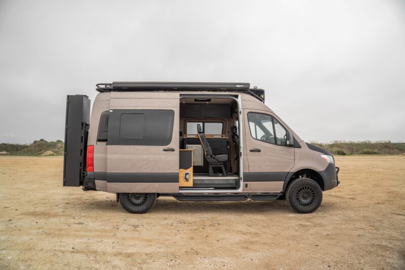 A beige Rogue Van Company camper van with its side door open, revealing the interior seating and storage, is parked on an empty, sandy area under a cloudy sky.