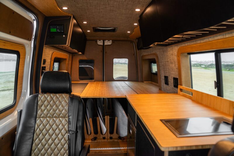 Interior of a modern camper van by Rogue Van Company featuring a leather driver's seat, built-in kitchenette with stove, wood counters, overhead cabinets, and a bed platform at the back.
