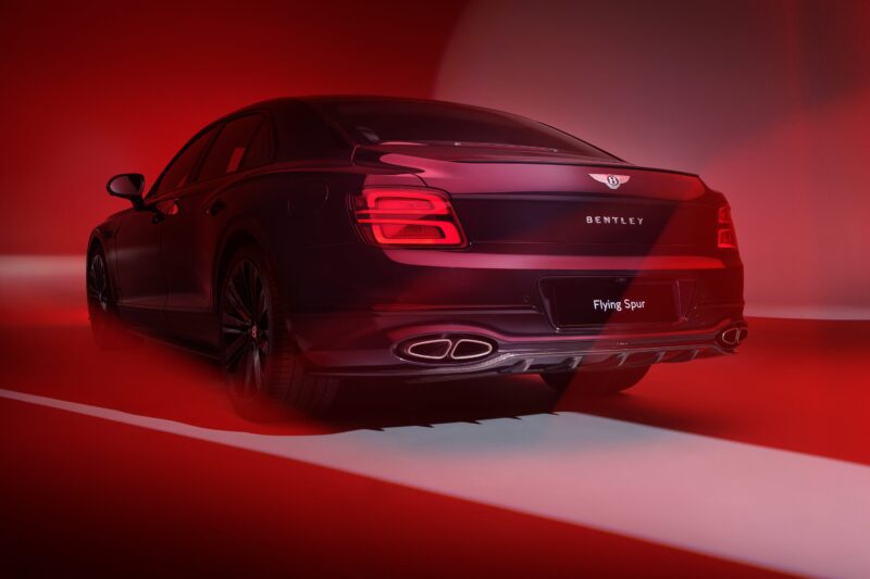 Rear view of a Bentley Flying Spur luxury sedan illuminated by dramatic red lighting, highlighting the taillights and dual exhaust pipes—a nod to Bentley Motors’ Akrapovič partnership, enhancing both design and exhaust note.