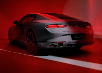 A sleek, silver Bentley Continental GT sports coupe viewed from the rear, illuminated by dramatic red lighting and shadows, showcases its unmistakable exhaust note—a testament to the Akrapovič partnership with Bentley Motors.