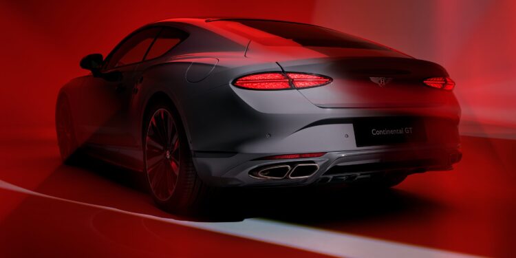 A sleek, silver Bentley Continental GT sports coupe viewed from the rear, illuminated by dramatic red lighting and shadows, showcases its unmistakable exhaust note—a testament to the Akrapovič partnership with Bentley Motors.