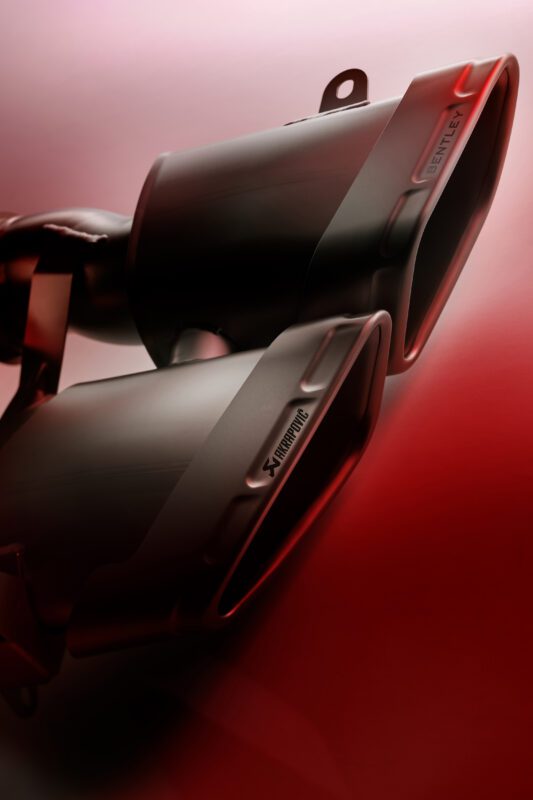 Close-up of dual black car exhaust tips with "Akrapovič" and "Bentley" labels, illuminated by red lighting—showcasing the thrilling exhaust note from the Akrapovič partnership with Bentley Motors.