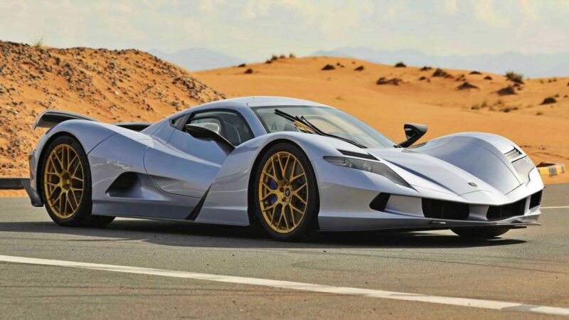 A silver sports car with gold rims, inspired by the most powerful cars of 2025, is parked on a paved road in a desert landscape under a partly cloudy sky.