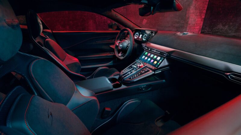 Interior view of the 2026 Aston Martin Vanquish S sports car featuring digital displays, leather seats, and a center console with a touchscreen interface, all illuminated by striking red ambient lighting.