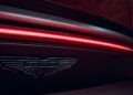 Close-up of an Aston Martin logo on the rear of a dark 2026 Aston Martin Vanquish S, with a red LED light strip and carbon fiber detailing above it.