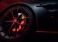 Close-up of a 2026 Aston Martin Vanquish S front wheel and brake disc, with red lighting reflecting off the rim and body, and an "S" badge visible on the side panel.