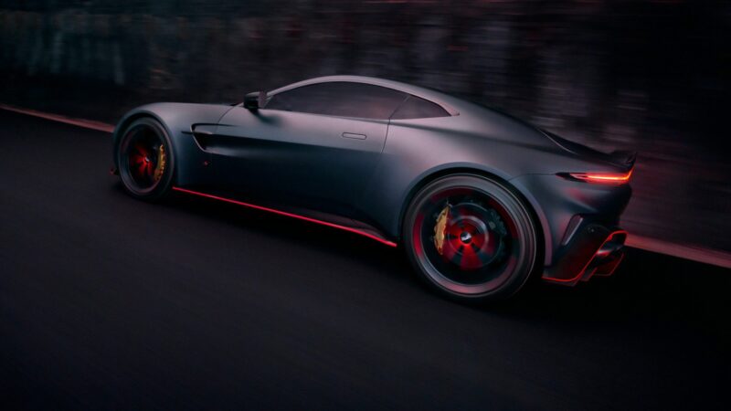 A matte black 2026 Aston Martin Vanquish S with red accents speeds down a dark road, creating a dramatic motion blur effect.