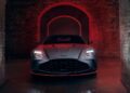 A matte gray 2026 Aston Martin Vanquish S with illuminated headlights is parked in a dimly lit brick tunnel accented by red lighting.