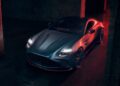 A matte grey 2026 Aston Martin Vanquish S with headlights on is parked in a dimly lit, industrial-style space featuring brick walls and red lighting.
